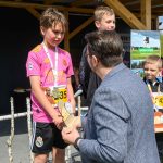 Na zdjęciu kilku chłopców stoi na podium. Jeden z nich, w różowej koszulce, otrzymuje nagrodę od mężczyzny. W tle widoczne zielone drzewa i zadaszona konstrukcja.