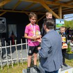 Na zdjęciu jest ceremonia wręczenia nagród z uśmiechniętymi dziećmi na podium. Dziecko w różowej koszulce trzyma trofeum. Tło to drewniana konstrukcja i kwiaty w doniczce.