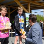 Na zdjęciu widać dwóch chłopców z medalami i nagrodami na tle drewnianej wiaty. Mężczyzna im gratuluje, a za nimi widać uczestników biegu. Tło zawiera niebo, drzewa i osoby w sportowych strojach.