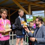 Chłopcy na podium z trofeami w otoczeniu dorosłego. Różne barwne ubrania, tło z plakatami. Słoneczny dzień na zewnątrz.
