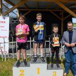 Trzech chłopców z medalami stoi na podium; liczby 1-3 są żółte. Po lewej białe tło z czerwonym napisem Mazowsze. Tło z drewnianą altaną i zieloną trawą.