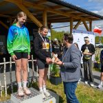 Na podium stoją dwie młode dziewczyny w sportowych strojach, jedna w zielono-niebieskiej bluzie. Mężczyzna w marynarce wręcza jej nagrodę. W tle widoczna jest polska flaga i grupa osób.