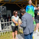 Na zdjęciu widać dekorację zwycięzców na podium. Kobieta odbiera nagrodę od mężczyzny na tle baneru z napisem Mazowsze. Tło jest ciemne z elementami drewna i zielenią trawy.