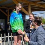 Na zdjęciu widać młodą dziewczynę na podium w zielono-niebieskiej bluzie. Otrzymuje nagrodę od mężczyzny. W tle drewniana altana i baner.