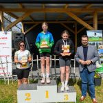 Na zdjęciu widać ceremonii wręczenia nagród na podium. Trzy osoby trzymają trofea, stojąc na tle drewnianego budynku. Jasne kolory ubrań kontrastują z ciemnym tłem.