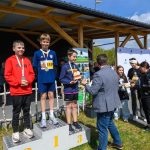 Trzech chłopców na podium w sportowych ubraniach z medalami na szyi. Tło drewniane z zadaszeniem, niebo zachmurzone. Obok wręczanie nagród.