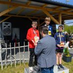 Trzech chłopców na podium otrzymuje medale i trofea. Mężczyzna wręcza trofeum. Tło stanowi drewniany budynek i transparenty.