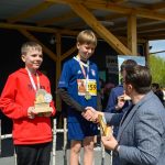 Na zdjęciu dwaj chłopcy otrzymują nagrody sportowe na podium. Jeden ma czerwoną bluzę, drugi niebieski strój sportowy. W tle drewniana konstrukcja i zieleń.