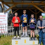Trzech chłopców stoi na podium z trofeami. Tło to drewniana altana z plakatami i zieloną trawą. Słoneczny dzień z niebieskim niebem.