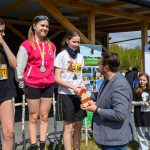 Na zdjęciu trzy kobiety na podium otrzymują medale. Tło stanowi drewniana konstrukcja i baner z logo Gminy Dobre. W tle widoczne są drzewa i kilka osób.
