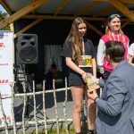 Mężczyzna wręcza nagrody trzem młodym kobietom podczas zawodów na scenie z logiem Mazowsze. Kobiety są uśmiechnięte i trzymają trofea. Tło stanowi drewniana konstrukcja i baner.