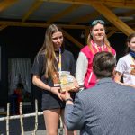 Trzy młode osoby stoją na podium, trzymając trofea. Tło to drewniana konstrukcja ze sceną i głośnikiem. Jest słoneczny dzień.