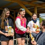 Trzy młode dziewczyny otrzymują nagrody na podium sportowym. Mają na sobie medale, jedna jest w różowej bluzie. W tle drewniane zadaszenie i zieleń.
