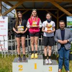Trzy młode kobiety stoją na podium, trzymają trofea. Mężczyzna po prawej w marynarce. Tło to drewniana altana i bannery.