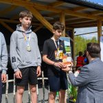 Trzech młodych sportowców z medalami stoi na podium, w tle drewniana konstrukcja. Jeden z nich odbiera nagrodę od mężczyzny. Słoneczny dzień, kilka osób obserwuje w tle.