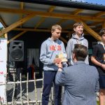 Trzech młodych sportowców na podium z medalami, w tle drewniana altana i baner Mazowsze. Kolory szary, niebieski i czerwony dominują na zdjęciu.