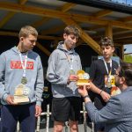 Trzech chłopców w bluzach i medalach trzyma trofea. Otrzymują je od mężczyzny w marynarce. W tle drewniana altana i zieleń.