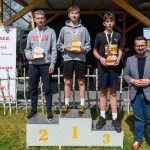 Trzech młodych sportowców stoi na podium, trzymając trofea. Obok nich stoi mężczyzna w marynarce. Tło to drewniana konstrukcja i banery.