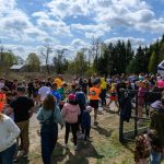 Grupa ludzi w kolorowych ubraniach czeka na start biegu. Niebieskie niebo z chmurami, w tle drzewa. Nad głowami różowo-biały łuk z napisem START.