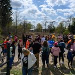 Grupa osób zebrana w parku, ubranych w kolorowe stroje sportowe. Tło stanowi las i niebo z chmurami. Ciepły, słoneczny dzień.