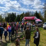 Na zdjęciu widnieje tłum ludzi na starcie biegu, z różnokolorowymi koszulkami. Nad nimi różowy łuk z napisem START. W tle drzewa i niebo z chmurami.