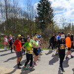 Grupa biegaczy w kolorowych strojach stoi na tle lasu. Dzień jest słoneczny, a niebo częściowo zachmurzone. Tło wypełniają drzew i zielona trawa.