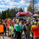 Grupa ludzi stoi przed różowo-białym łukiem z napisem START. Uczestnicy są ubrani w kolorowe stroje sportowe. Tło stanowi zielone drzewo i niebo z białymi chmurami.