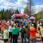 Grupa ludzi zgromadzona przy różowo-białym łuku startowym, otoczona zielenią. Uczestnicy ubrani w jaskrawe stroje sportowe. Niebo z niebieskimi i białymi chmurami.