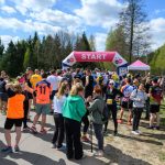Grupa ludzi w kolorowych ubraniach czeka pod różowym bannerem START. W tle drzewa i błękitne, pochmurne niebo. Atmosfera sportowa i radosna.