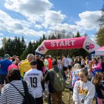 Na zdjęciu grupa uczestników biegu gromadzi się pod różowym łukiem startowym. Niebo jest niebieskie z białymi chmurami. W tle widoczne są drzewa i biały namiot.