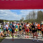 Grupa biegaczy stoi na linii startu pod różowym łukiem. W tle są drzewa i niebieskie niebo. Po lewej widać biało-niebieski namiot.