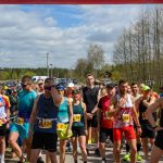 Biegacze na linii startu, kolorowe stroje, słoneczny dzień. W tle las i niebo z chmurami. Grupa ludzi przygotowuje się do startu.