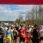 Grupa biegaczy stoi na linii startu biegu na świeżym powietrzu. Ubrani w kolorowe stroje sportowe, otoczeni zielenią lasu. Słoneczny dzień, na niebie widoczne są chmury.