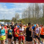Na zdjęciu grupa biegaczy w kolorowych strojach stoi na linii startu na zewnątrz. Tło stanowią drzewa i lekko pochmurne niebo. Widoczne są numery startowe i kibice w oddali.