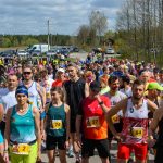 Biegacze w kolorowych strojach czekają na start na tle lasu i nieba. Uliczka pełna ludzi. Słoneczny dzień, dużo zieleni.