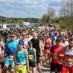 Grupa osób w kolorowych ubraniach czeka na start biegu. Za nimi widać drzewa i kilka zaparkowanych aut. Nad nimi rozciąga się niebieskie niebo z chmurami.