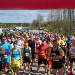 Grupa biegaczy na starcie maratonu, kolorowe stroje i numery startowe. Tło to las i parking z samochodami. Pogoda jest słoneczna.