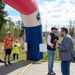 Na zdjęciu widać start biegu, uczestnicy przygotowują się. Nad nimi wisi duży, kolorowy łuk startowy. W tle zielone drzewa i niebo.
