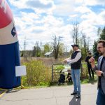 Mężczyzna z mikrofonem stoi obok niebiesko-czerwonego łuku startowego. W tle widać zielone drzewa i błękitne niebo. Dzień jest słoneczny.