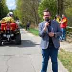 Mężczyzna w okularach przemawia do mikrofonu na tle zielonych drzew. Obok przejeżdża quad straży pożarnej. Grupa ludzi obserwuje z boku.