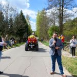 Na zdjęciu grupa ludzi na drodze w parku, mężczyzna mówi do mikrofonu. Obok czerwony quad, w tle drzewa i niebo. Dzień jest słoneczny.