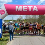 Biegacze stoją pod różowo-białym łukiem z napisem META na tle nieba i drzew. Wydarzenie sportowe odbywa się na asfalcie. Po lewej stronie widoczny zegar czasu.