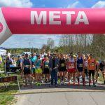Biegacze stoją na starcie pod różowo-niebieskim łukiem z napisem META. W tle widoczny jest las i kilka chmur na błękitnym niebie.