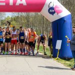 Grupa biegaczy stoi na linii startu pod różowym napisem META. W tle widać drzewa, a z boku stoi mężczyzna z trąbką. Obok bramy startowej siedzą dzieci.