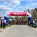 Na zdjęciu grupa biegaczy startuje spod różowo-niebieskiego namiotu z napisem META. W tle są drzewa i błękitne niebo z chmurami. Obserwatorzy stoją po bokach trasy.
