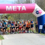 Grupa biegaczy startuje spod różowo-białego balonu z napisem META. Tło stanowią drzewa i niebo. Uczestnicy są ubrani kolorowo.