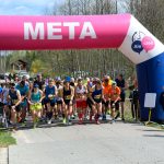 Grupa biegaczy startuje pod różowoniebieskim balonowym łukiem z napisem META. W tle zalesiony krajobraz. Kibice i fotografowie obserwują start.