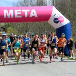 Grupa biegaczy startuje spod różowo-niebieskiego balonu z napisem META na tle zalesionego krajobrazu. Na zdjęciu dominuje ruch i energia, a uczestnicy noszą kolorowe stroje sportowe. W tle widoczni są kibice i sędzia z mikrofon