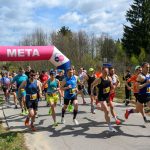 Biegacze startujący z linii mety pod różowo-białym łukiem. Uczestnicy w kolorowych strojach, wokół zielone drzewa. Dzień słoneczny, tłum kibiców.