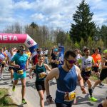 Biegacze przekraczają różowo-białą metę, a za nimi widać las i błękitne niebo. Kolorowe stroje uczestników. Spokojna atmosfera.